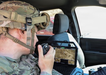 A U.S. Army soldier identified by name tape as 'HARTMAN' is seated inside a military vehicle operating communications equipment. The soldier is wearing MultiCam (OCP) combat uniform with a tactical helmet equipped with a mounted device, and is speaking into a handheld radio while surrounded by multiple laptop computers displaying mapping and surveillance data. The setup suggests a mobile command or reconnaissance operation.