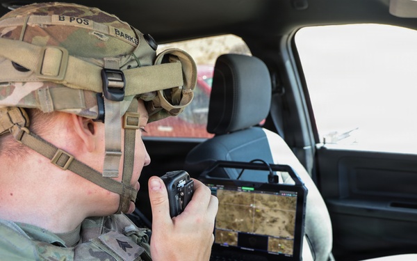 A U.S. Army soldier identified by name tape as 'HARTMAN' is seated inside a military vehicle operating communications equipment. The soldier is wearing MultiCam (OCP) combat uniform with a tactical helmet equipped with a mounted device, and is speaking into a handheld radio while surrounded by multiple laptop computers displaying mapping and surveillance data. The setup suggests a mobile command or reconnaissance operation.