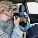 A U.S. Army soldier identified by name tape as 'HARTMAN' is seated inside a military vehicle operating communications equipment. The soldier is wearing MultiCam (OCP) combat uniform with a tactical helmet equipped with a mounted device, and is speaking into a handheld radio while surrounded by multiple laptop computers displaying mapping and surveillance data. The setup suggests a mobile command or reconnaissance operation.