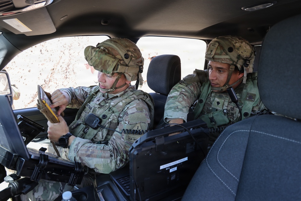 Two U.S. Army soldiers in full combat gear, including OCP (Operational Camouflage Pattern) uniforms and helmets, are seated inside a military vehicle reviewing information. The soldier on the left appears to be consulting a small notebook or card while the soldier on the right observes a mounted tactical computer or display console. The setting suggests a field training exercise or operational mission briefing.