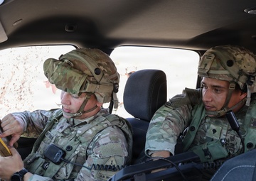 Two U.S. Army soldiers in full combat gear, including OCP (Operational Camouflage Pattern) uniforms and helmets, are seated inside a military vehicle reviewing information. The soldier on the left appears to be consulting a small notebook or card while the soldier on the right observes a mounted tactical computer or display console. The setting suggests a field training exercise or operational mission briefing.