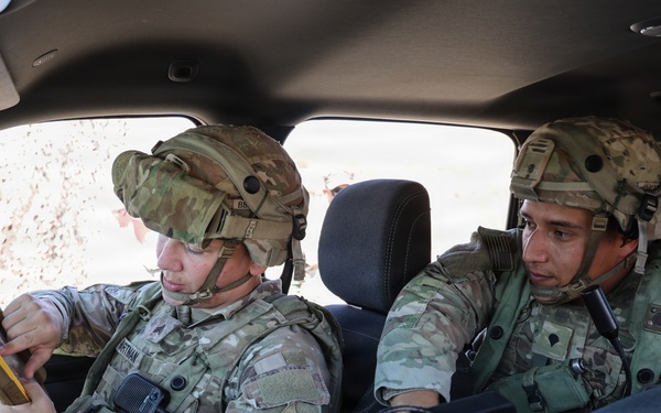 Two U.S. Army soldiers in full combat gear, including OCP (Operational Camouflage Pattern) uniforms and helmets, are seated inside a military vehicle reviewing information. The soldier on the left appears to be consulting a small notebook or card while the soldier on the right observes a mounted tactical computer or display console. The setting suggests a field training exercise or operational mission briefing.