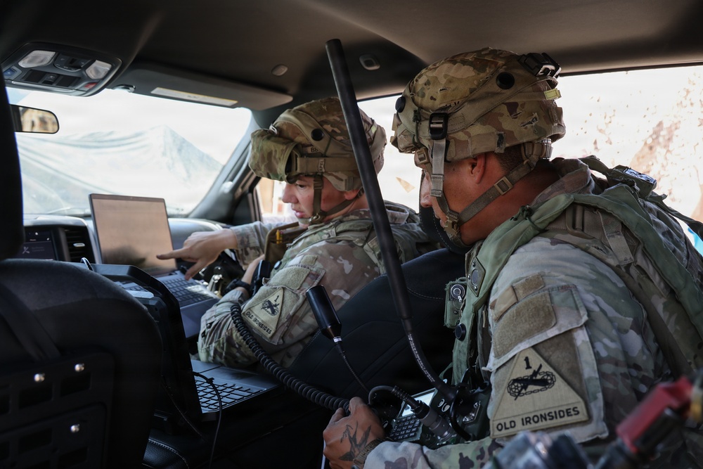 Two U.S. Army soldiers in full combat gear are seated inside a military vehicle, reviewing information on a laptop computer. Both wear MultiCam (OCP) uniforms and helmets with tactical accessories. The soldier on the right displays a '1st Armored Division - Old Ironsides' shoulder patch.