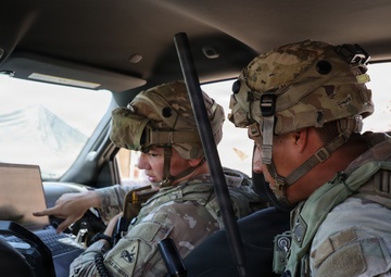 Two U.S. Army soldiers in full combat gear are seated inside a military vehicle, reviewing information on a laptop computer. Both wear MultiCam (OCP) uniforms and helmets with tactical accessories. The soldier on the right displays a '1st Armored Division - Old Ironsides' shoulder patch.
