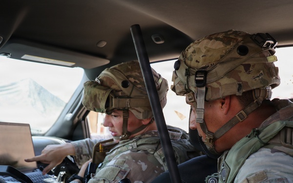Two U.S. Army soldiers in full combat gear are seated inside a military vehicle, reviewing information on a laptop computer. Both wear MultiCam (OCP) uniforms and helmets with tactical accessories. The soldier on the right displays a '1st Armored Division - Old Ironsides' shoulder patch.