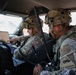Two U.S. Army soldiers in full combat gear are seated inside a military vehicle, reviewing information on a laptop computer. Both wear MultiCam (OCP) uniforms and helmets with tactical accessories. The soldier on the right displays a '1st Armored Division - Old Ironsides' shoulder patch.