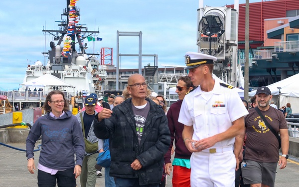 A group of civilians and a U.S. Navy officer in dress whites walk along a waterfront pier or dock area. A naval vessel decorated with signal flags is visible in the background, suggesting a public event such as Fleet Week or a Navy open house. The setting appears to be an urban waterfront, likely in the Pacific Northwest based on visible signage and architecture.