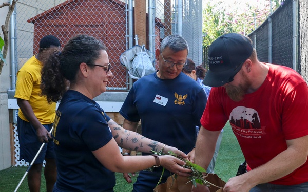 A group of volunteers are engaged in outdoor cleanup or landscaping work in what appears to be a fenced yard or animal shelter area. A woman in a navy shirt is placing plant trimmings into a large brown paper bag held by a man in a red t-shirt. Additional volunteers are visible in the background, one using a rake on artificial turf.