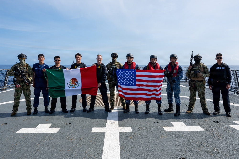 A group of approximately eleven military and law enforcement personnel pose for a group photo on the flight deck of a naval vessel at sea. They are holding the flags of Mexico and the United States, suggesting a joint bilateral maritime operation or exercise. Personnel are wearing a mix of uniforms including U.S. Coast Guard (USCG), Mexican Navy (Marina), and tactical gear.