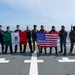 A group of approximately eleven military and law enforcement personnel pose for a group photo on the flight deck of a naval vessel at sea. They are holding the flags of Mexico and the United States, suggesting a joint bilateral maritime operation or exercise. Personnel are wearing a mix of uniforms including U.S. Coast Guard (USCG), Mexican Navy (Marina), and tactical gear.
