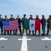 A group of approximately eleven military and law enforcement personnel pose for a group photo on what appears to be a ship's helicopter deck. They are holding two national flags — the United States flag and the Mexican flag — suggesting a joint bilateral military or law enforcement operation. Personnel are wearing a mix of uniforms including U.S. Coast Guard, U.S. Customs and Border Protection, camouflage tactical gear, and Mexican Navy uniforms.