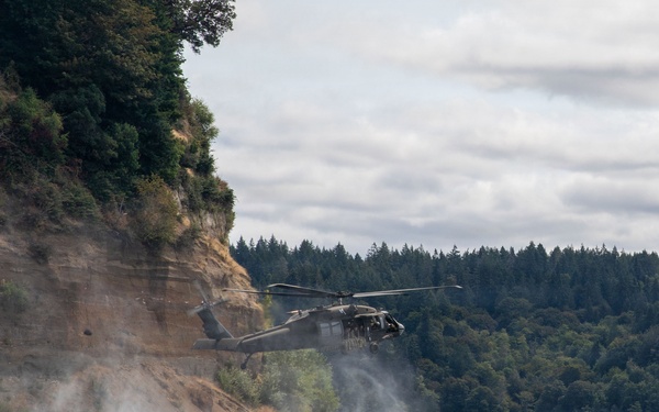 A military helicopter, consistent with a UH-60 Black Hawk, is hovering at low altitude near a rocky cliff face over a body of water. The rotor wash is visibly disturbing the water surface and kicking up mist or dust near the shoreline. The setting appears to be a forested coastal or inland waterway environment under overcast skies.