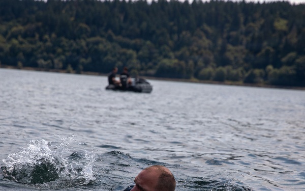 A person is swimming through open water, appearing to be in full or partial clothing, suggesting a military or training exercise. In the background, a small inflatable boat with at least one occupant is visible on the water. The setting appears to be a large lake or bay surrounded by dense coniferous forest.