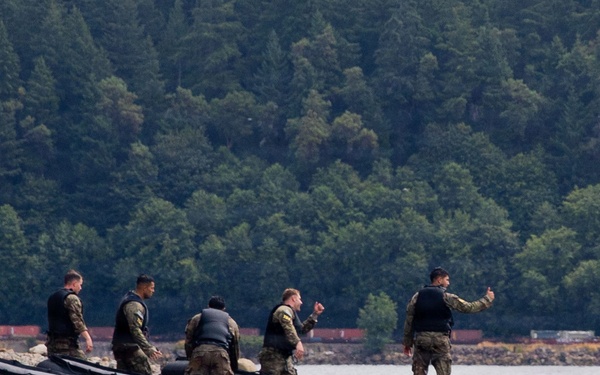 Five military personnel in camouflage uniforms and tactical vests are standing on a large black inflatable pontoon or raft structure on a body of water. One individual at the front appears to be giving hand signals or directing the group. The background shows a rocky shoreline and a dense coniferous forest, suggesting a Pacific Northwest or similar temperate coastal environment.