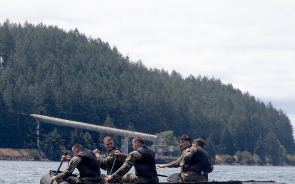 A group of military personnel in camouflage uniforms and wetsuits are paddling a black inflatable rubber raiding craft (IBS/Zodiac-type boat) on open water. The scene appears to be a military training exercise, with a forested shoreline and what appears to be a military installation visible in the background. The overcast conditions and tactical gear suggest a realistic operational training scenario.