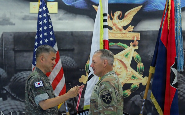 A military ceremony is taking place at the 2nd Infantry Division Hall of Fame, where a Republic of Korea Army officer is presenting a medal or decoration to a U.S. Army officer in ACU/OCP uniform. The backdrop features the 2nd Infantry Division insignia and a historical combat mural. A third soldier in Korean Army uniform stands at attention to the right.