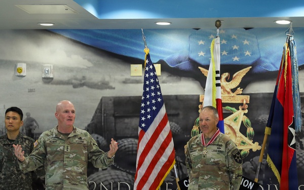 A military ceremony is taking place in front of a 2nd Infantry Division 'Hell on Wheels' backdrop, likely at Camp Red Cloud or a similar installation in South Korea. Two U.S. Army officers are prominently featured, one speaking and one wearing a medal/decoration, alongside a Republic of Korea Army soldier. The setting includes the American flag and a unit guidon, suggesting a formal awards or change of command ceremony.