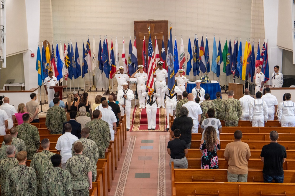 A formal U.S. Navy military ceremony is taking place inside what appears to be a chapel or auditorium. Officers in white dress uniforms are positioned at the front near a color guard holding flags, while attendees in both military uniforms and civilian attire stand in pews. The backdrop features numerous U.S. state and territorial flags arranged in a semicircle.