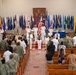 A formal U.S. Navy military ceremony is taking place inside what appears to be a chapel or auditorium. Officers in white dress uniforms are positioned at the front near a color guard holding flags, while attendees in both military uniforms and civilian attire stand in pews. The backdrop features numerous U.S. state and territorial flags arranged in a semicircle.