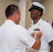 A military promotion or award ceremony is taking place indoors, where one individual in white Navy dress uniform is pinning an insignia or device onto another uniformed Navy senior enlisted member. The recipient, wearing a combination cover (officer/CPO-style cap) with gold insignia, displays multiple service ribbons and qualification badges on his uniform. Multiple flags are visible in the background, indicating a formal ceremonial setting.