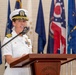A U.S. Navy female officer in white dress uniform is speaking at a podium during a formal military ceremony. The podium bears a 'Naval Station' seal/emblem. Multiple U.S. state flags are displayed in the background.