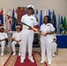 Two U.S. Navy personnel in white dress uniforms are standing and jointly holding a decorated ceremonial oar or paddle during what appears to be a formal military ceremony. Two additional naval officers are seated in the background observing the event. The setting features numerous U.S. state and territorial flags displayed on stands behind the participants.