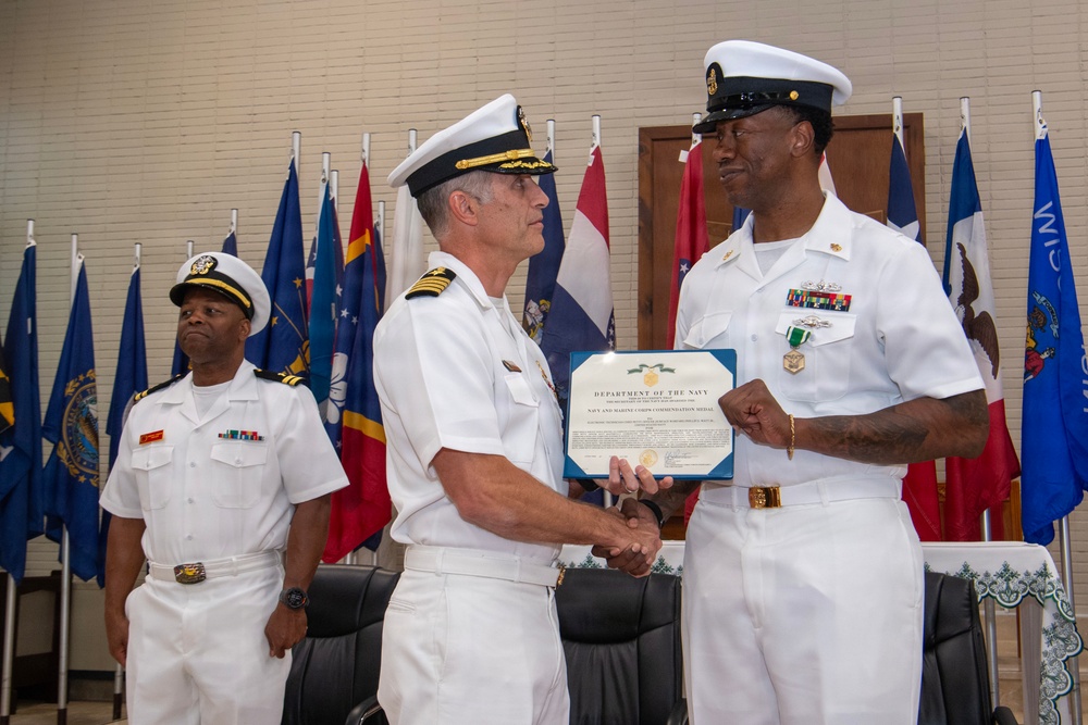 A formal U.S. Navy award ceremony is taking place in which an officer is presenting a Department of the Navy certificate and medal to a senior enlisted sailor. The recipient is wearing a Navy and Marine Corps Commendation Medal on his white uniform. A third Navy officer stands at attention in the background.