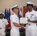 A formal U.S. Navy award ceremony is taking place in which an officer is presenting a Department of the Navy certificate and medal to a senior enlisted sailor. The recipient is wearing a Navy and Marine Corps Commendation Medal on his white uniform. A third Navy officer stands at attention in the background.