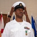 A U.S. Navy Chief Petty Officer in white dress uniform is adjusting his combination cover (cap) during what appears to be a formal military ceremony. He is wearing multiple service ribbons, a warfare device badge, and a green Navy achievement or commendation medal around his neck. Multiple flags are visible in the background, consistent with a ceremonial hall setting.