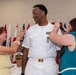 A U.S. Navy military promotion or retirement ceremony is taking place indoors. A tall service member in dress white uniform is having insignia or devices pinned onto his collar/shoulders by two civilian women, likely family members. Multiple flags and a uniformed officer in the background indicate a formal military setting.