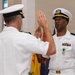 Two U.S. Navy officers in white service uniforms are engaged in what appears to be a formal military ceremony, likely an oath of office or promotion ceremony. Both officers have their right hands raised in a swearing-in gesture. The setting includes multiple state and territorial flags in the background, suggesting an official military installation or ceremonial hall.