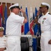 A formal U.S. Navy military ceremony is taking place, likely a change of command or retirement ceremony. Two naval officers in white dress uniforms are exchanging salutes in the foreground, while a third officer seated in the background observes with a smile. Multiple U.S. state and organizational flags are displayed prominently behind the participants.