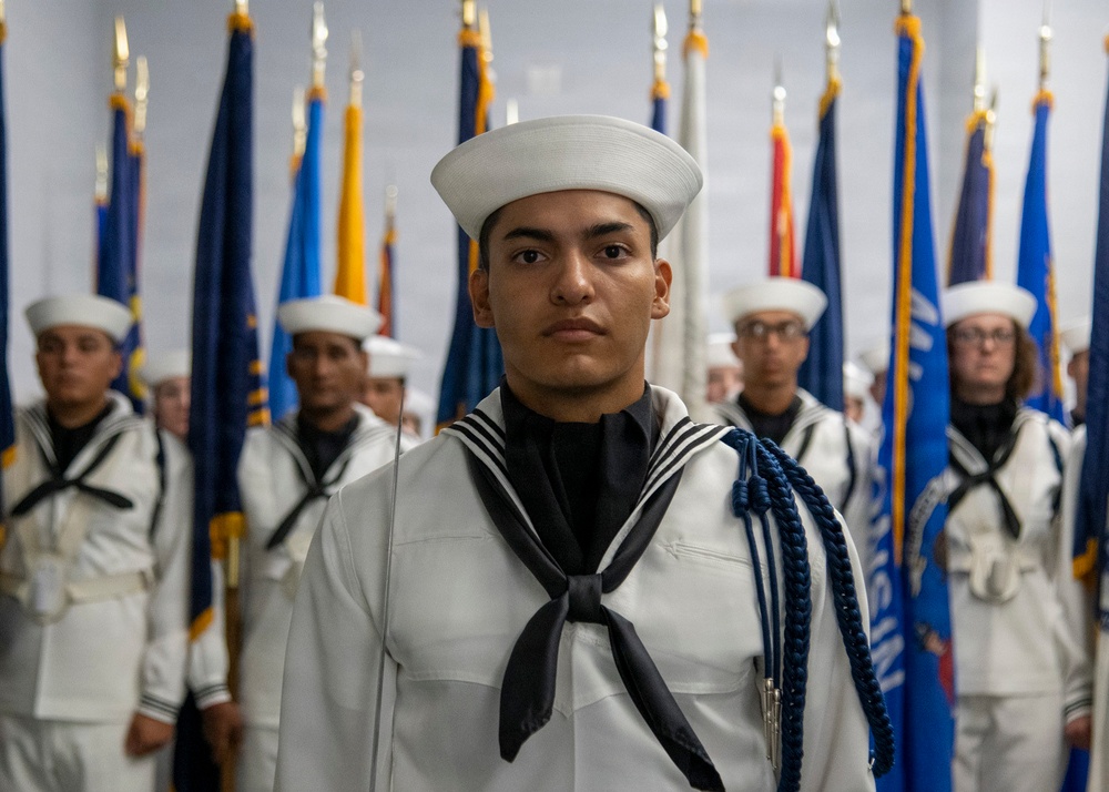 A group of U.S. Navy sailors in dress white uniforms stand at attention in a formal ceremonial formation. The foreground sailor wears a distinctive blue aiguillette (braided cord), indicating a ceremonial or honor guard role. Multiple state and organizational flags are displayed in the background, suggesting an official military ceremony or graduation event.