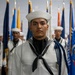 A group of U.S. Navy sailors in dress white uniforms stand at attention in a formal ceremonial formation. The foreground sailor wears a distinctive blue aiguillette (braided cord), indicating a ceremonial or honor guard role. Multiple state and organizational flags are displayed in the background, suggesting an official military ceremony or graduation event.