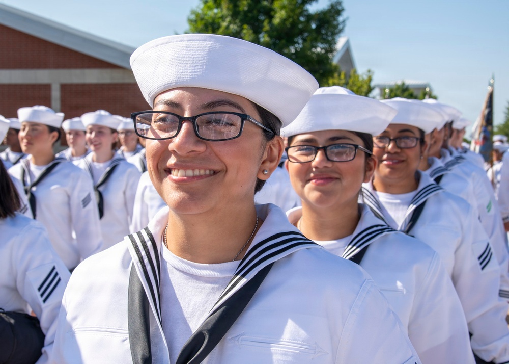 A group of individuals dressed in white U.S. Navy enlisted dress white uniforms, including the distinctive white sailor caps (dixie cups), are assembled outdoors in formation. The foreground features a smiling young woman wearing black-rimmed glasses and a navy neckerchief, with several similarly uniformed personnel visible behind her. The setting appears to be a formal military event or ceremony, likely a graduation or pass-in-review.