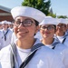 A group of individuals dressed in white U.S. Navy enlisted dress white uniforms, including the distinctive white sailor caps (dixie cups), are assembled outdoors in formation. The foreground features a smiling young woman wearing black-rimmed glasses and a navy neckerchief, with several similarly uniformed personnel visible behind her. The setting appears to be a formal military event or ceremony, likely a graduation or pass-in-review.