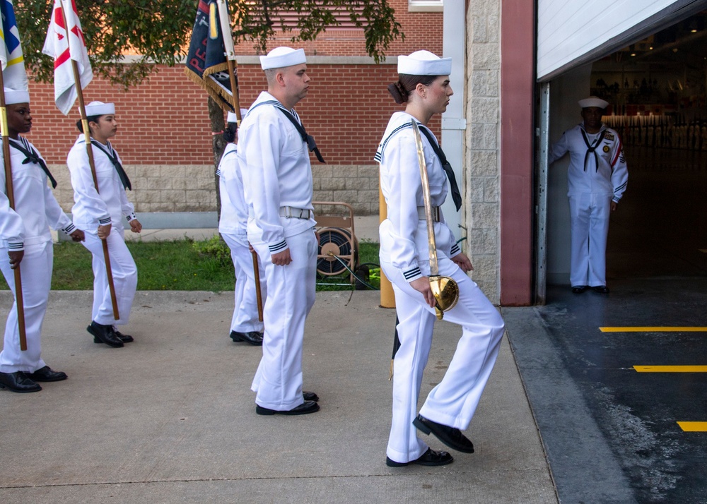 A group of U.S. Navy personnel in white dress uniforms are marching in a ceremonial formation, likely a color guard or parade drill. The lead sailor carries a ceremonial sword, while others behind carry flags and guidons. A supervising sailor in white uniform observes from a doorway to the right.