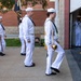 A group of U.S. Navy personnel in white dress uniforms are marching in a ceremonial formation, likely a color guard or parade drill. The lead sailor carries a ceremonial sword, while others behind carry flags and guidons. A supervising sailor in white uniform observes from a doorway to the right.