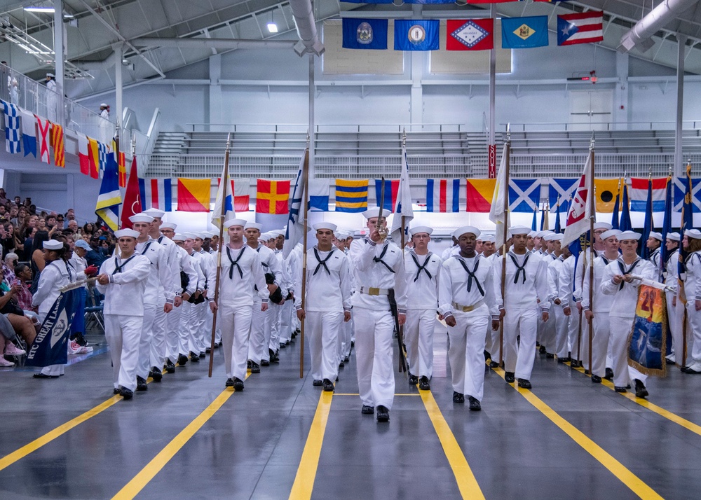 A U.S. Navy recruit graduation ceremony (Pass in Review) is taking place inside a large indoor drill hall at RTC Great Lakes, Illinois. Rows of recruits dressed in white dress uniforms are marching in formation while carrying guidon flags and rifles. Spectators are seated in bleachers along the sides, and numerous naval signal flags and U.S. state flags are displayed throughout the facility.