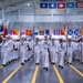 A U.S. Navy recruit graduation ceremony (Pass in Review) is taking place inside a large indoor drill hall at RTC Great Lakes, Illinois. Rows of recruits dressed in white dress uniforms are marching in formation while carrying guidon flags and rifles. Spectators are seated in bleachers along the sides, and numerous naval signal flags and U.S. state flags are displayed throughout the facility.