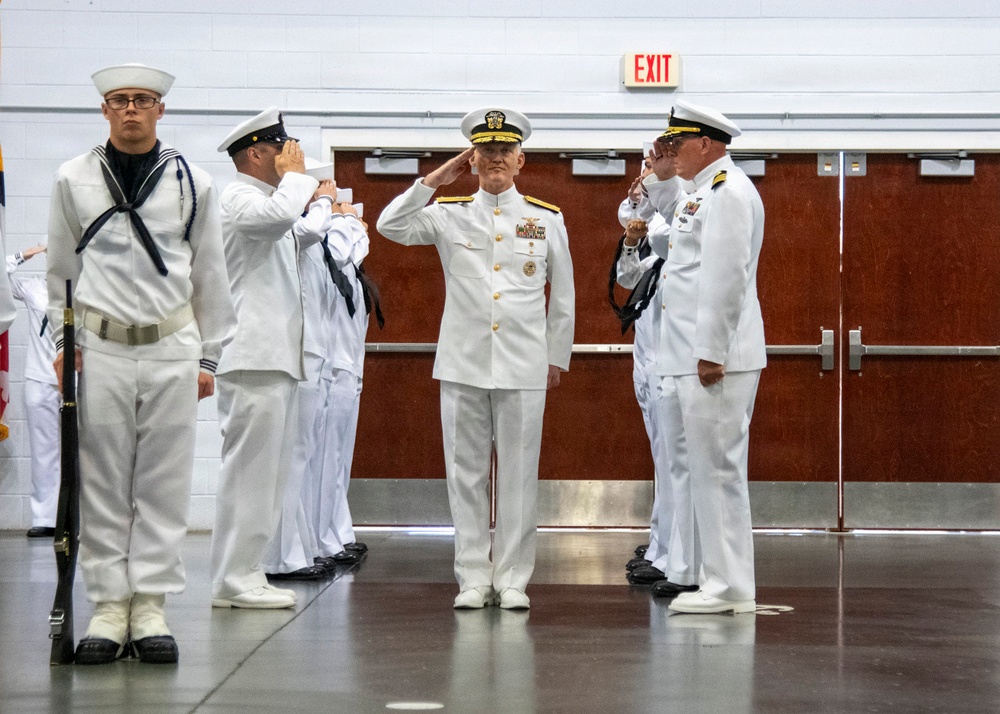 A formal U.S. Navy military ceremony is taking place indoors, likely a change of command or official ceremony. Multiple naval personnel in white dress uniforms are rendering salutes, while one enlisted sailor stands at attention holding a rifle as part of a color guard. The setting appears to be a gymnasium or large indoor facility.