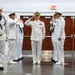 A formal U.S. Navy military ceremony is taking place indoors, likely a change of command or official ceremony. Multiple naval personnel in white dress uniforms are rendering salutes, while one enlisted sailor stands at attention holding a rifle as part of a color guard. The setting appears to be a gymnasium or large indoor facility.