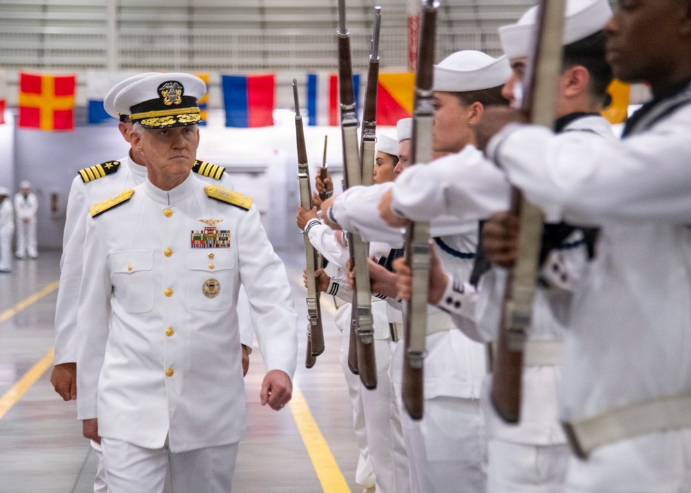 A senior U.S. Navy flag officer in Service Dress White uniform walks through a ceremonial sideboy or honor guard formation inside what appears to be a large hangar or drill hall. Sailors in white uniforms hold rifles at present arms, forming a corridor for the officer's passage. The setting and formation suggest a formal military ceremony such as a change of command, retirement, or commissioning event.