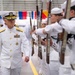 A senior U.S. Navy flag officer in Service Dress White uniform walks through a ceremonial sideboy or honor guard formation inside what appears to be a large hangar or drill hall. Sailors in white uniforms hold rifles at present arms, forming a corridor for the officer's passage. The setting and formation suggest a formal military ceremony such as a change of command, retirement, or commissioning event.