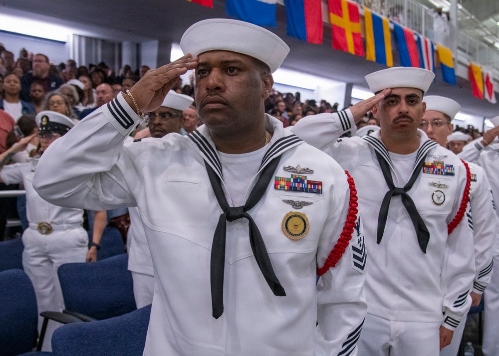 U.S. Navy sailors in dress white uniforms are rendering a salute during a formal military ceremony. The foreground sailor displays multiple service ribbons, a command medallion, and aviation warfare insignia. A large audience of civilians and military personnel is visible in the background beneath a row of international flags.