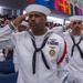 U.S. Navy sailors in dress white uniforms are rendering a salute during a formal military ceremony. The foreground sailor displays multiple service ribbons, a command medallion, and aviation warfare insignia. A large audience of civilians and military personnel is visible in the background beneath a row of international flags.
