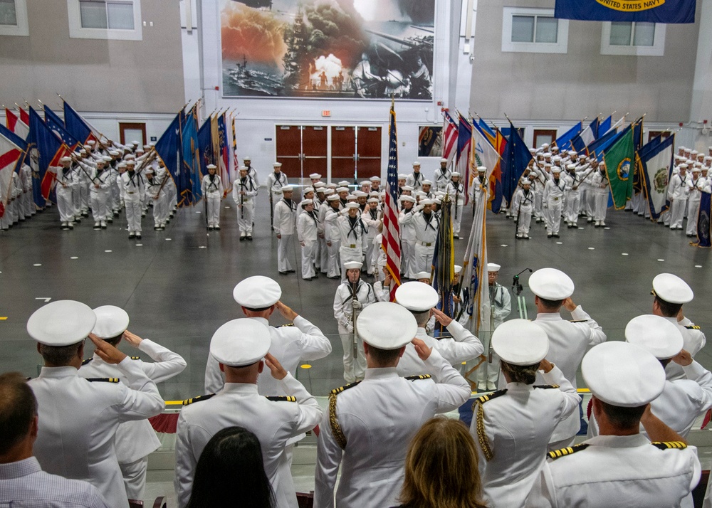 A formal United States Navy military ceremony is taking place inside a large indoor hall. Naval officers in white dress uniforms are saluting a color guard presenting the American flag and state flags. A large mural depicting what appears to be the attack on Pearl Harbor is prominently displayed on the back wall.
