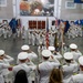 A formal United States Navy military ceremony is taking place inside a large indoor hall. Naval officers in white dress uniforms are saluting a color guard presenting the American flag and state flags. A large mural depicting what appears to be the attack on Pearl Harbor is prominently displayed on the back wall.