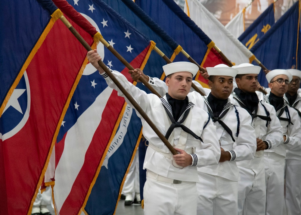 A group of U.S. Navy sailors in dress white uniforms are participating in a formal color guard ceremony, carrying multiple flags including state and military flags. The sailors are standing in a disciplined line formation, each holding flag staffs at an angle. The setting appears to be an indoor military ceremony or parade.