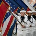 A group of U.S. Navy sailors in dress white uniforms are participating in a formal color guard ceremony, carrying multiple flags including state and military flags. The sailors are standing in a disciplined line formation, each holding flag staffs at an angle. The setting appears to be an indoor military ceremony or parade.